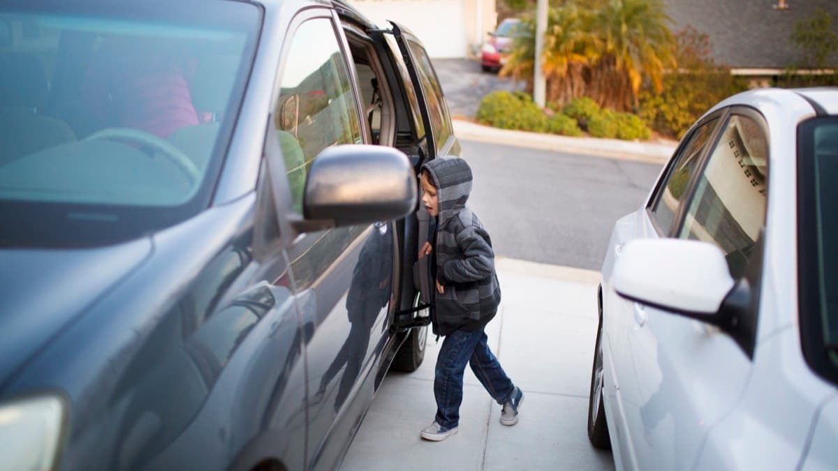 A small boy walking between a minivan and a parked car in a driveway
