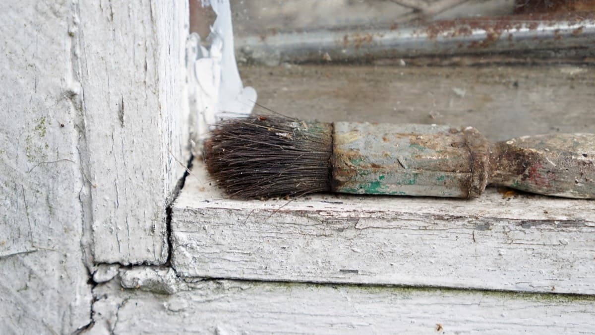 Cracked, flaking white paint on an old wooden window frame, with a worn paintbrush on the sill
