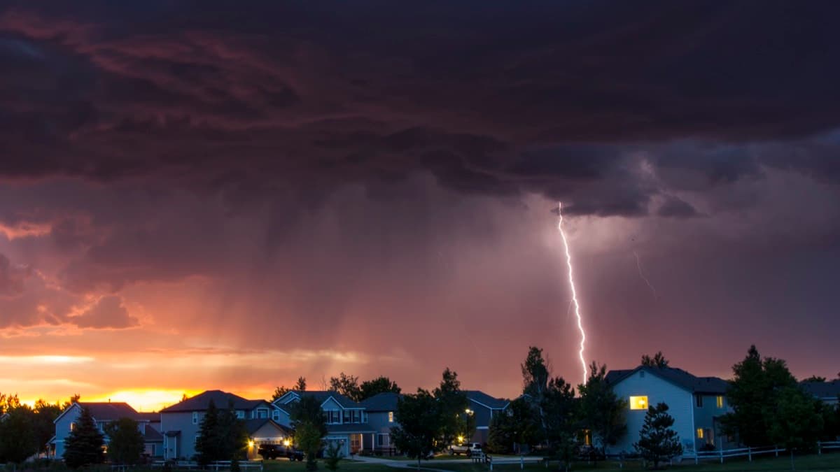 A lightning bolt striking beyond a row of suburban homes under a stormy sunset sky