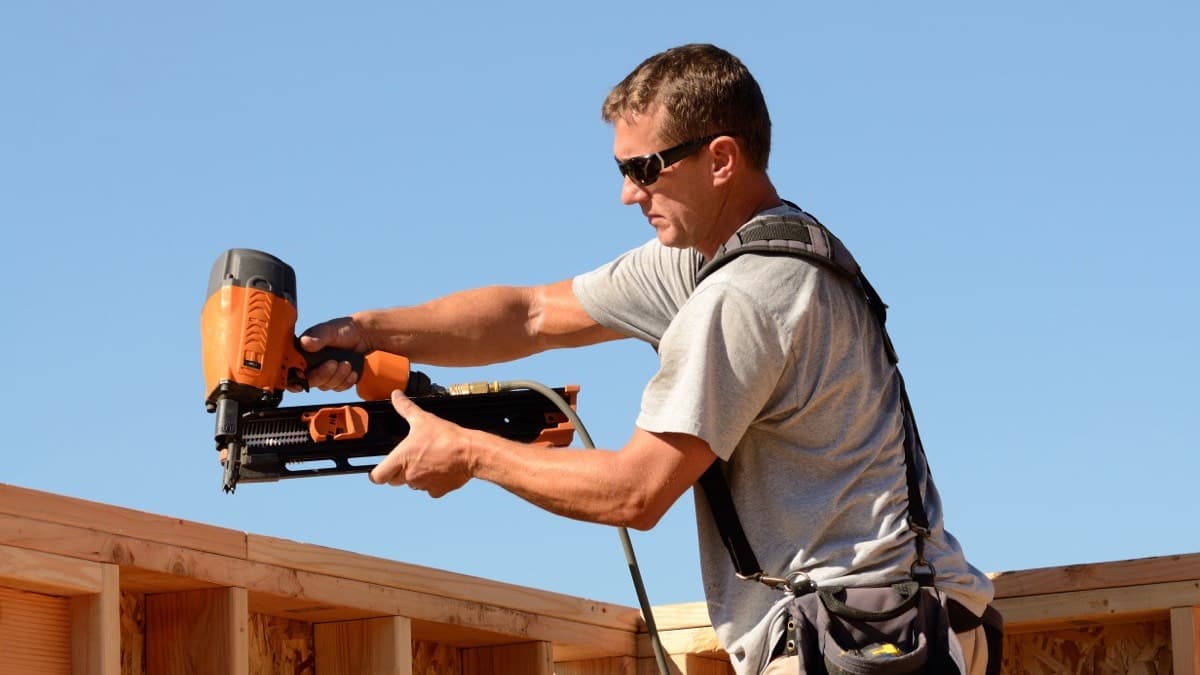 A framer in safety glasses driving nails with a pneumatic nail gun on new wall framing