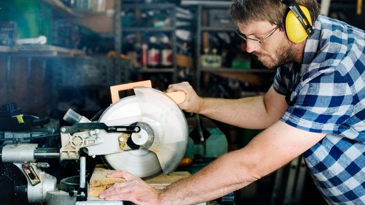 A carpenter in yellow ear muffs and safety glasses cutting a board on a miter saw in a workshop