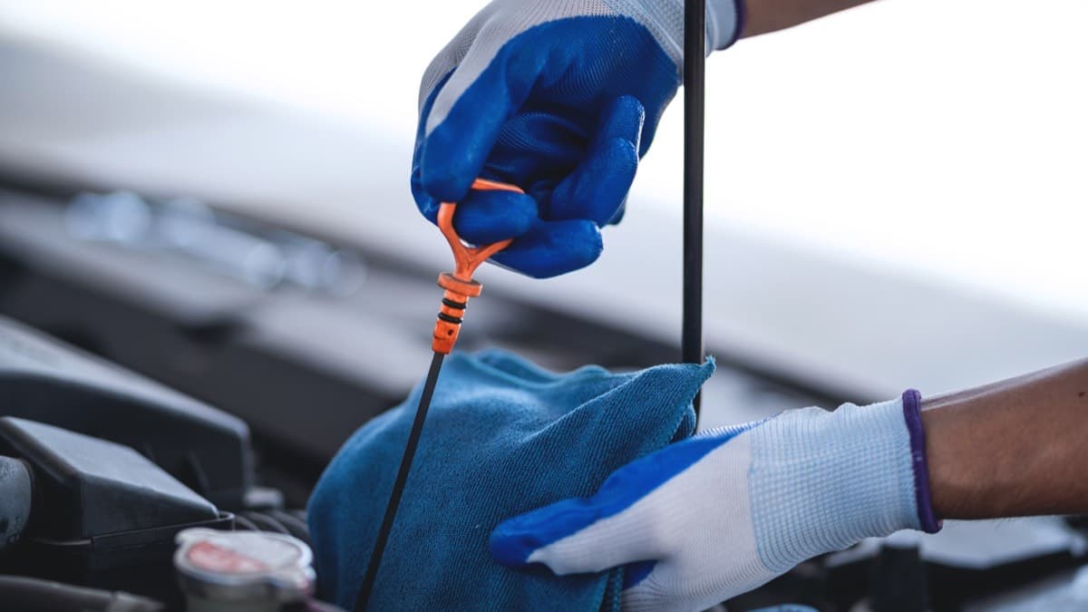 Gloved hands pulling an engine oil dipstick and wiping it on a blue rag under an open hood