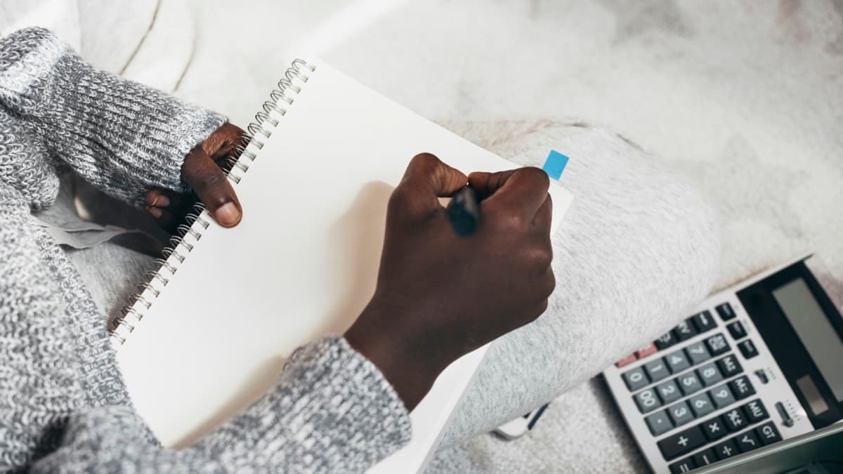 A person writing in a spiral notebook beside a desk calculator