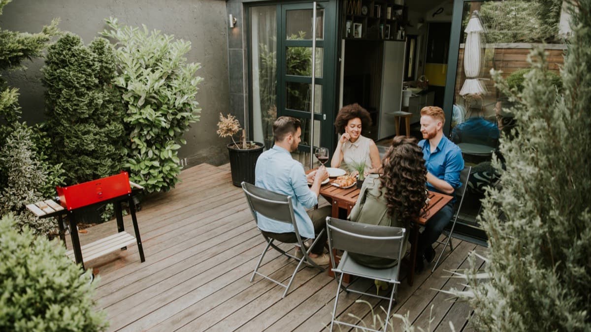 Four guests eating dinner at a table on a backyard deck beside a red grill