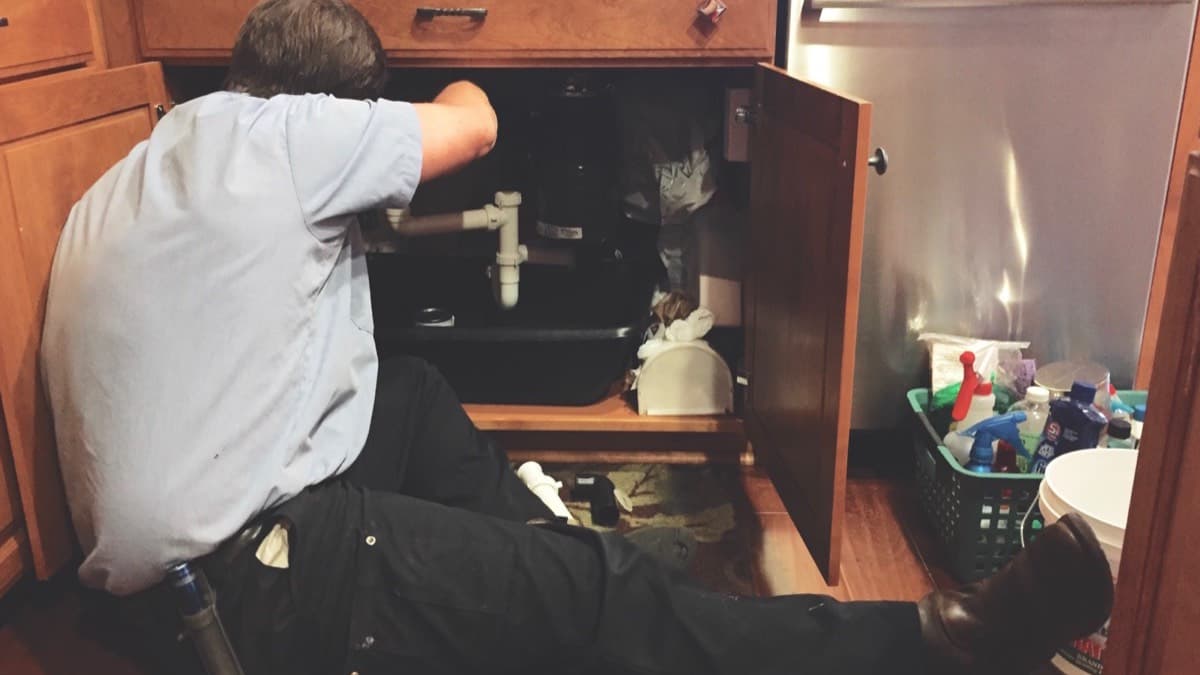 A maintenance worker lying on a kitchen floor working on the pipes under a sink cabinet