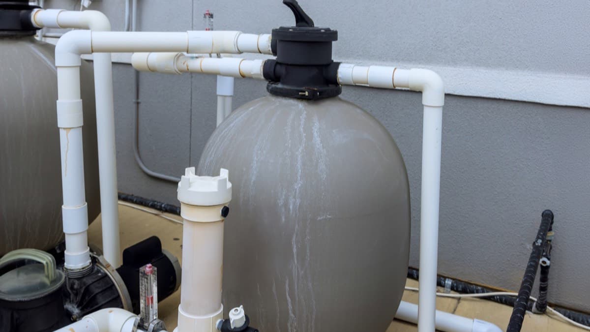 A pool sand filter, pump and white PVC pipework on an equipment pad beside a house wall