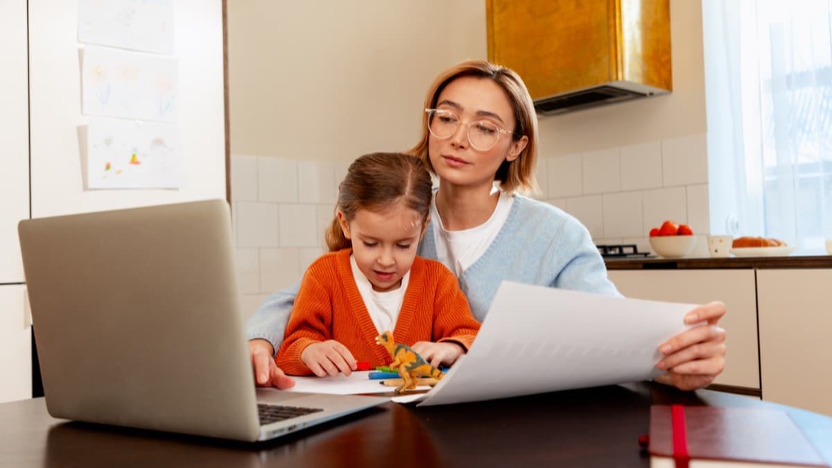 A woman reading a paper at a laptop with a young girl on her lap