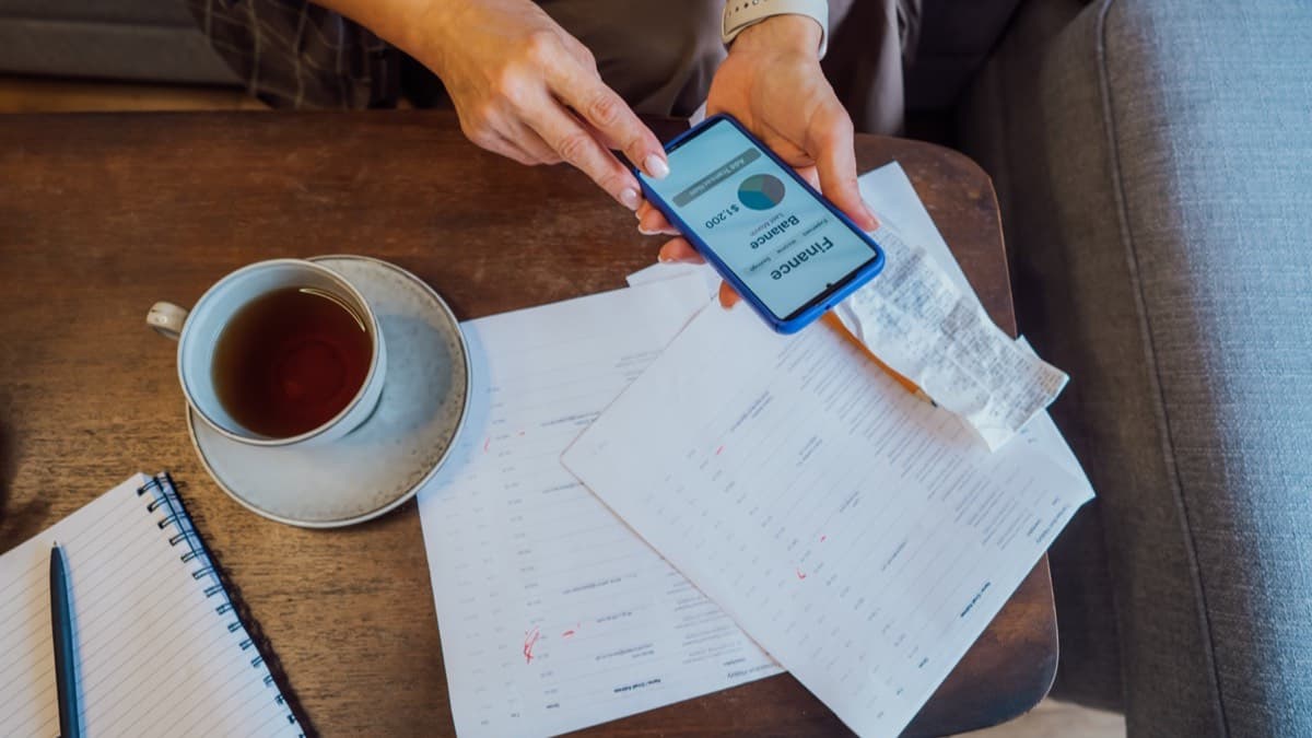 Hands holding a phone with a banking app over printed statements marked in red pen