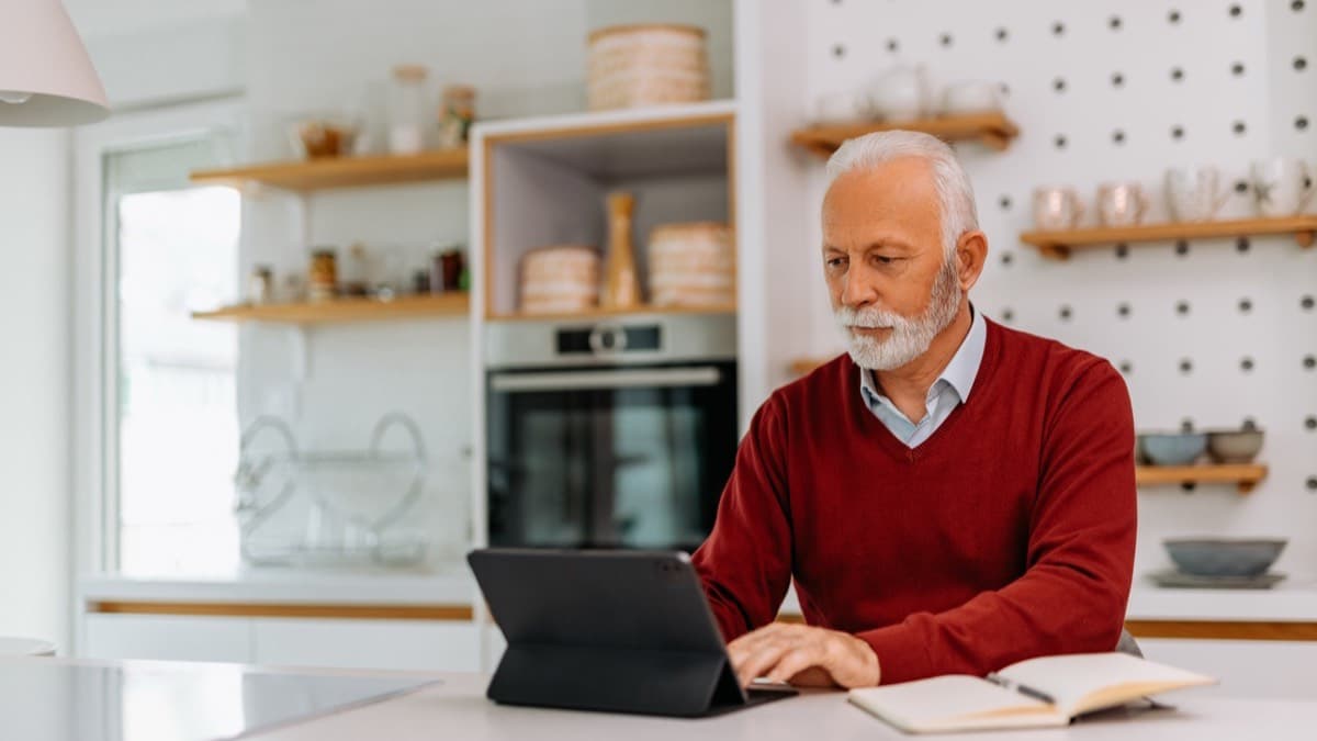 An older man typing on a tablet at a kitchen counter beside an open notebook