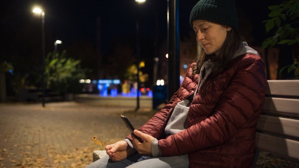 A person sitting alone on a park bench at night, looking at a phone