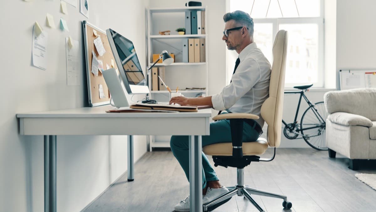 A person sitting upright in an office chair at a desk with a monitor, in a home office beside a window