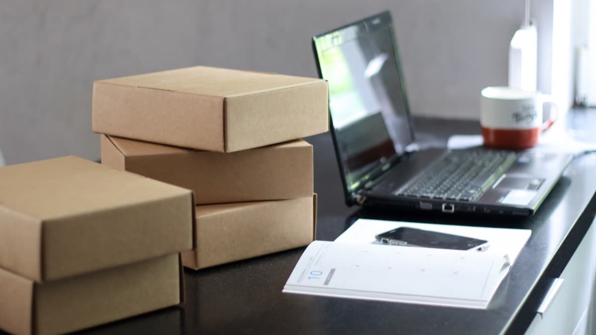 Stacked cardboard delivery boxes on a desk beside an open laptop, a planner and a phone