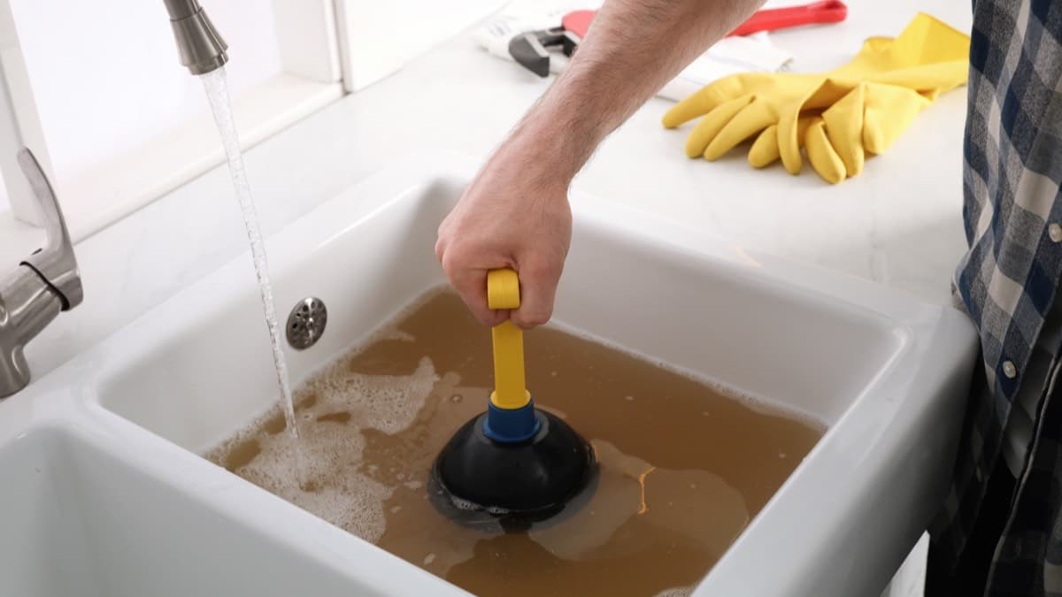 A hand working a plunger in a sink full of murky standing water, with rubber gloves and a wrench on the counter