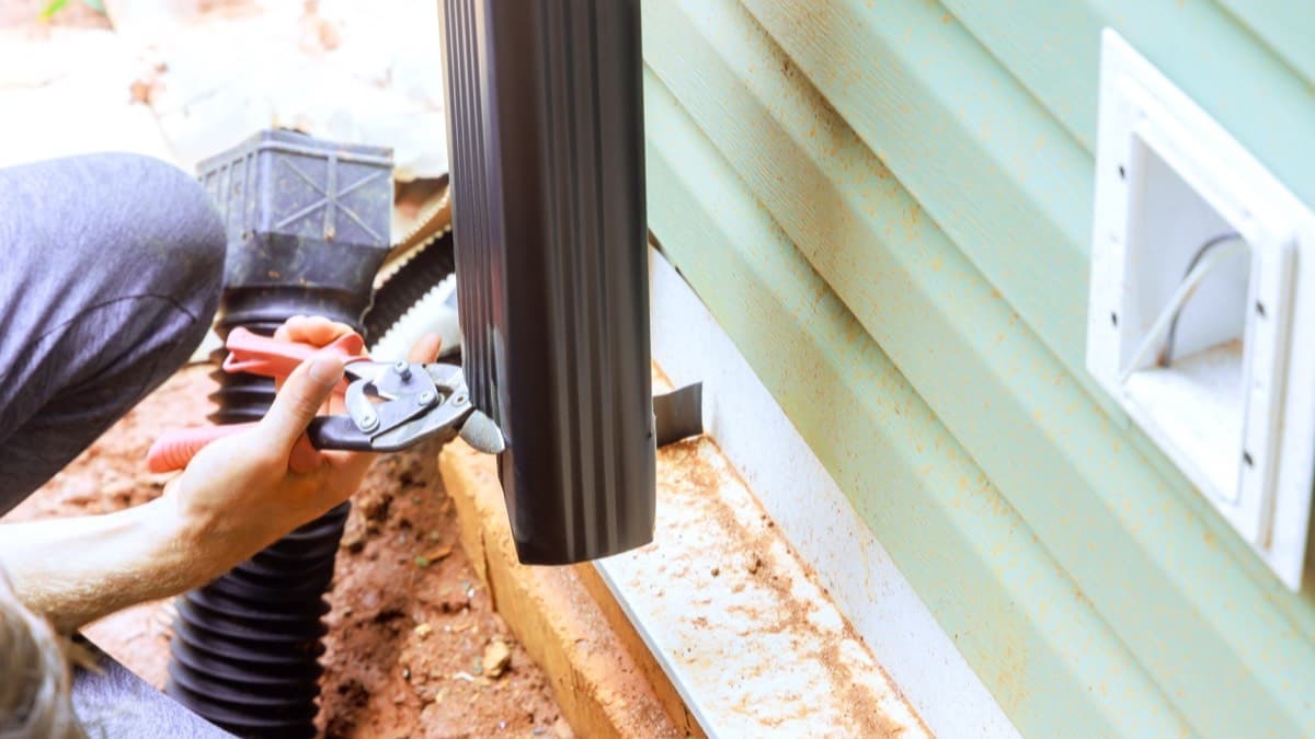 A person fitting a downspout into a corrugated drain line that carries rainwater away from a sided house
