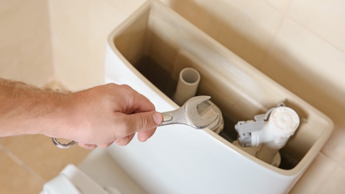 A hand with a wrench over an open toilet tank showing the overflow tube and fill valve
