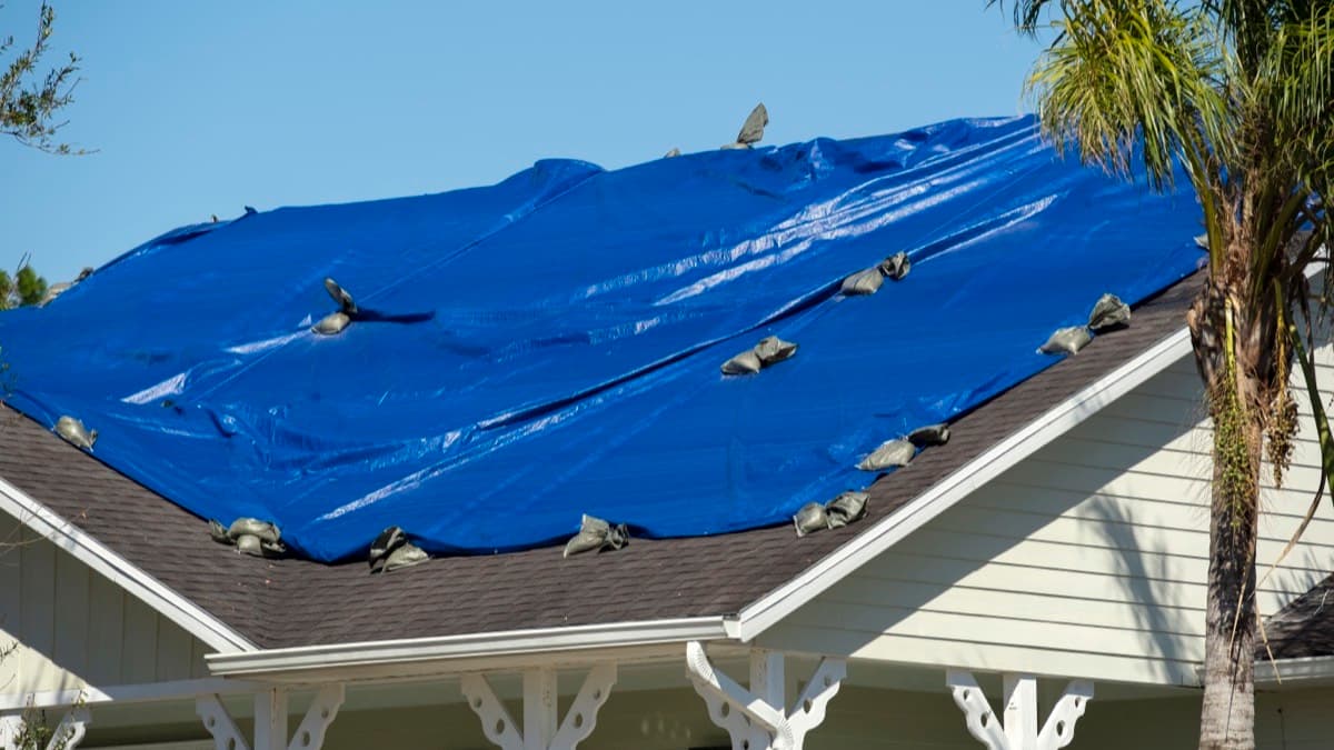 A blue tarp held down with sandbags over a damaged shingle roof after a storm