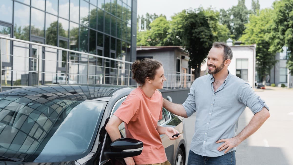 A parent talking with a teenager holding car keys beside a parked car