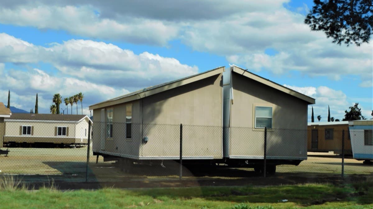 A manufactured home section on its chassis at a lot behind a chain-link fence
