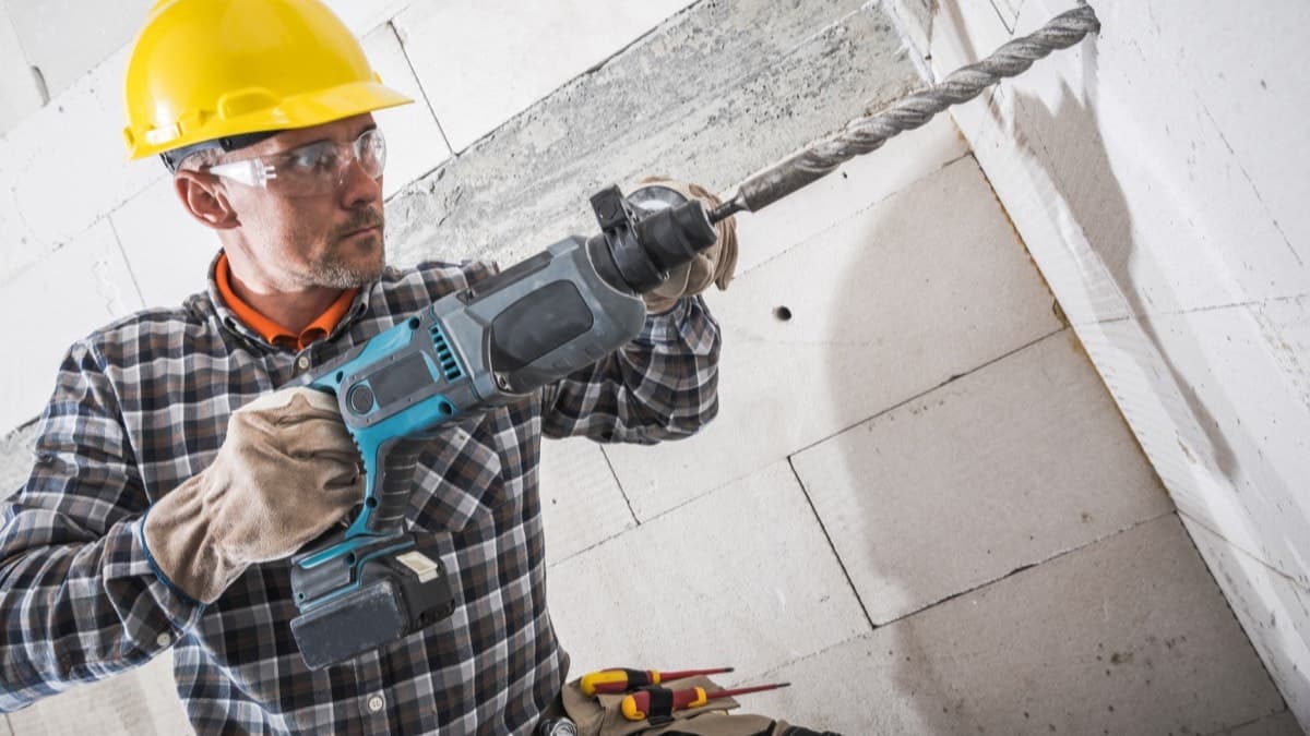 A worker in a hard hat, safety glasses and gloves drilling into a wall with a rotary hammer