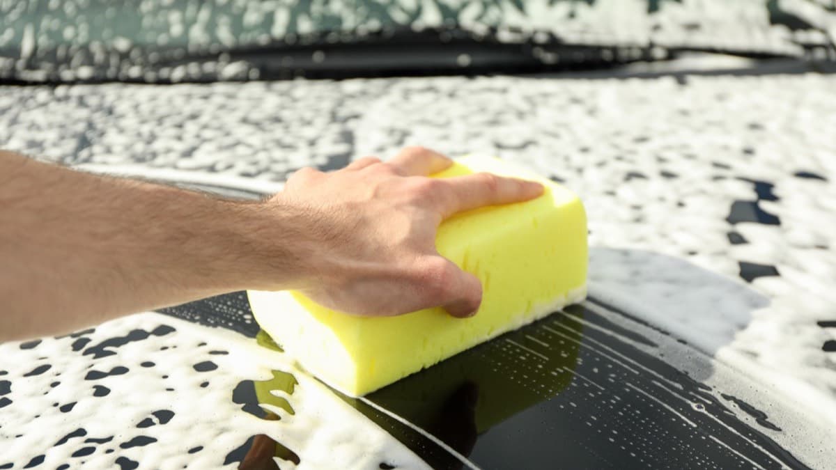 A hand scrubbing a soapy car hood with a yellow sponge