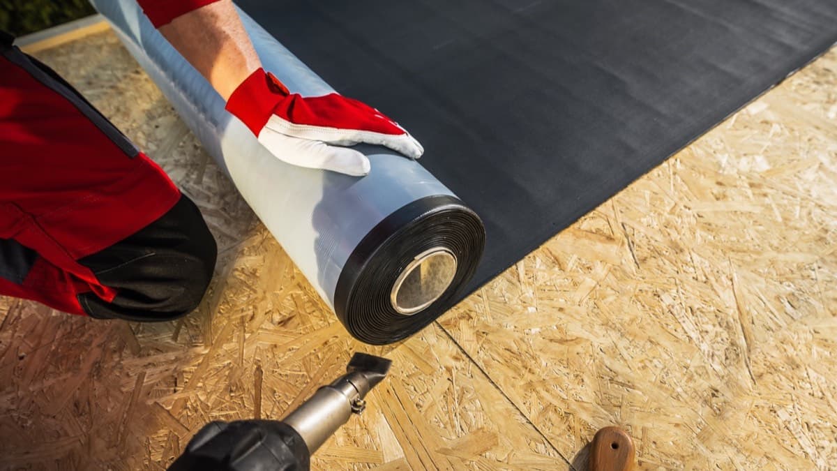 A roofer in work gloves rolling a sealing membrane across a bare wood roof deck