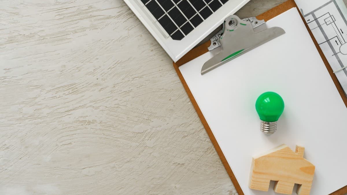 A clipboard with a green light bulb and a small wooden house beside a solar panel and floor plans