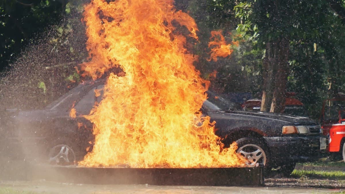Flames engulfing the front of a parked dark sedan as water spray hits the fire