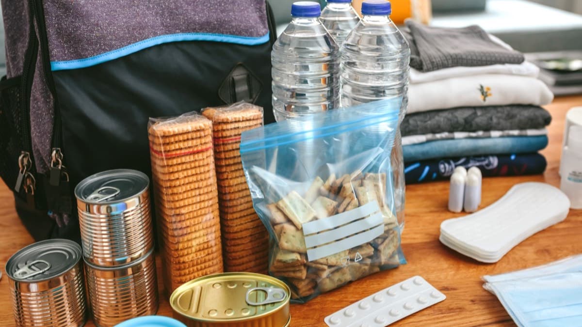 Bottled water, canned food, crackers, medicine and face masks laid out on a table beside a backpack
