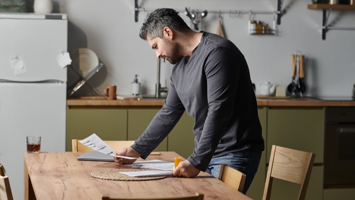 A person leaning over a kitchen table to read papers spread beside a laptop