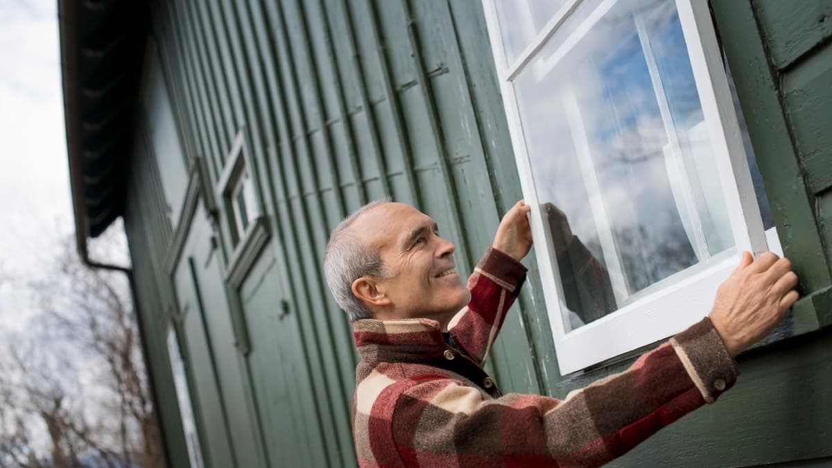 A person fitting a new white-framed window into the green wood siding of a house