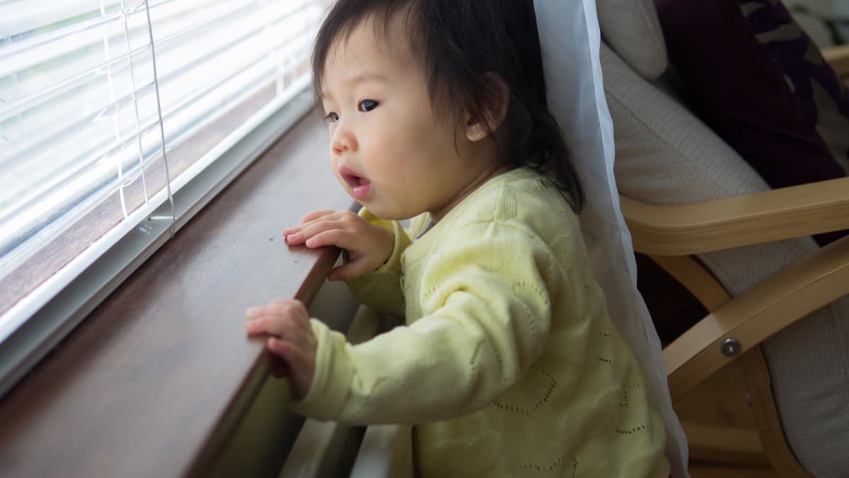 A toddler standing at a low window sill holding on and looking out past the blinds